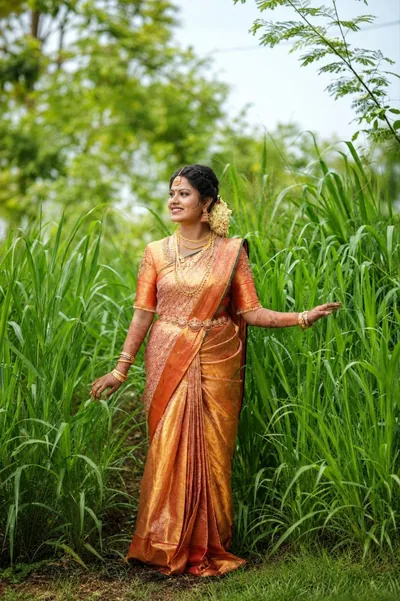 Bride in a gold-orange silk saree standing in tall green grass